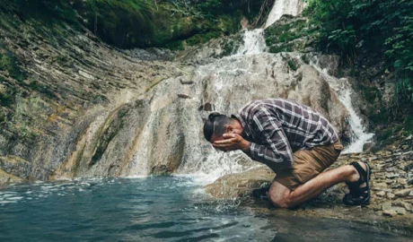 Hiker crouching at the edge of a waterfall pool, splashing water on his face to cool down