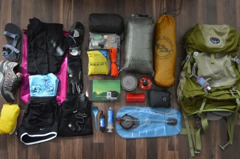 Hiking gear laid out on the floor: backpack, tent, sleeping bag, clothing, first aid kit, water bladder and cooking items