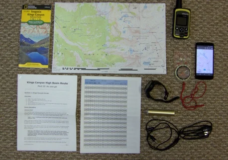 Navigation gear laid out for a hike: printed maps, route notes, GPS unit, compass, phone and charging cables