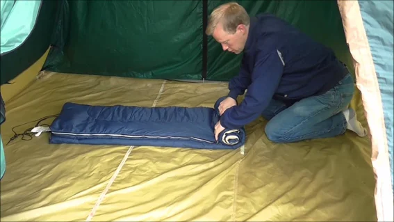 Man kneeling in a tent, tightly rolling up a sleeping bag to save space in his pack