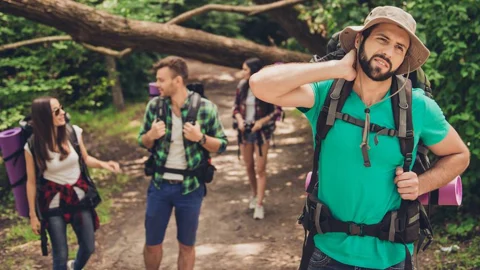Hiker wearing a heavy backpack rubbing his sore neck on a forest trail, with friends walking behind