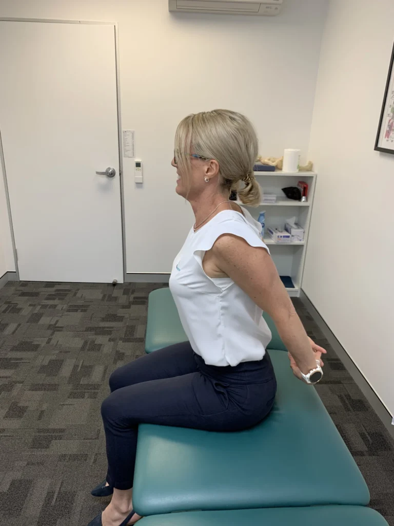 Woman seated on a treatment bed, hands clasped behind her back and shoulders drawn back to open the chest