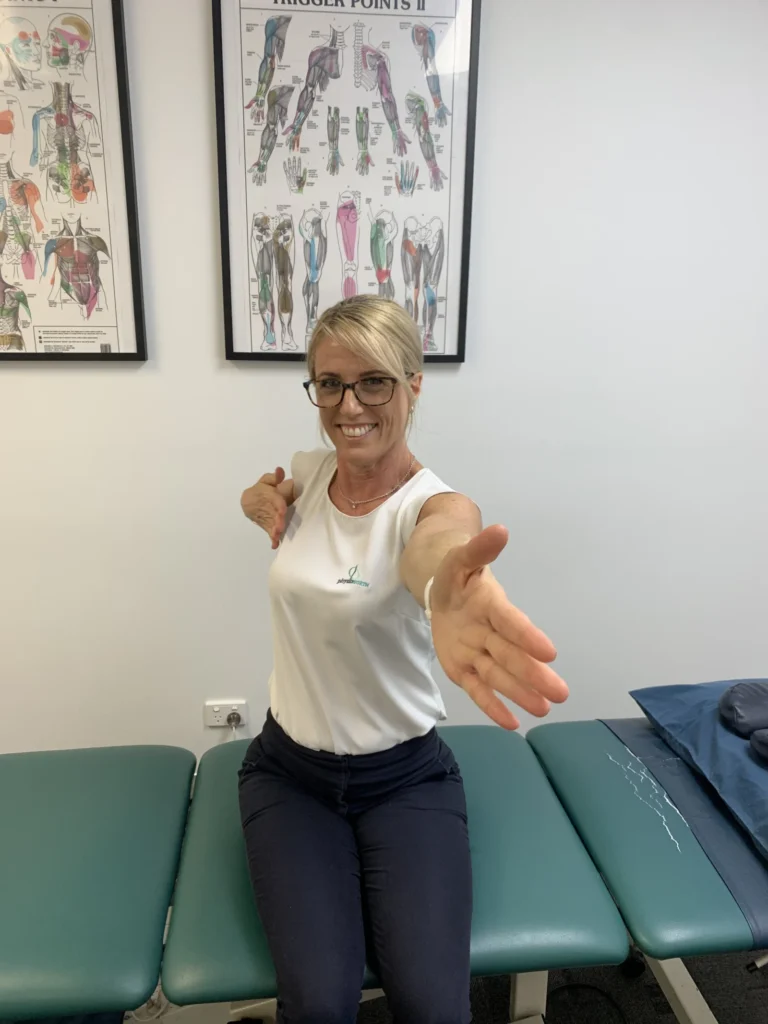 Woman seated on a treatment bed, arms out wide with palms forward to stretch the chest and front of the shoulders