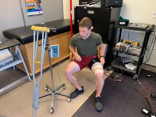 Man seated on a chair with a blood flow restriction cuff on his thigh, connected to a pressure unit, crutches nearby