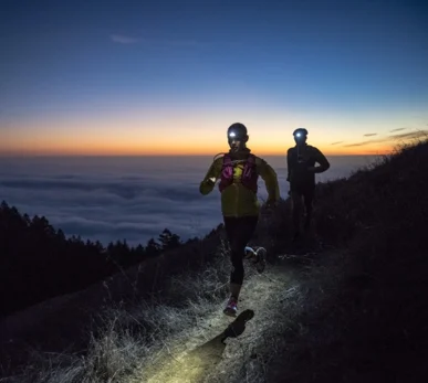 Two trail runners with head torches running along a ridge at dusk, above a sea of cloud