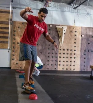 Man balancing on one foot on an unstable wobble board in a gym, arms out for balance