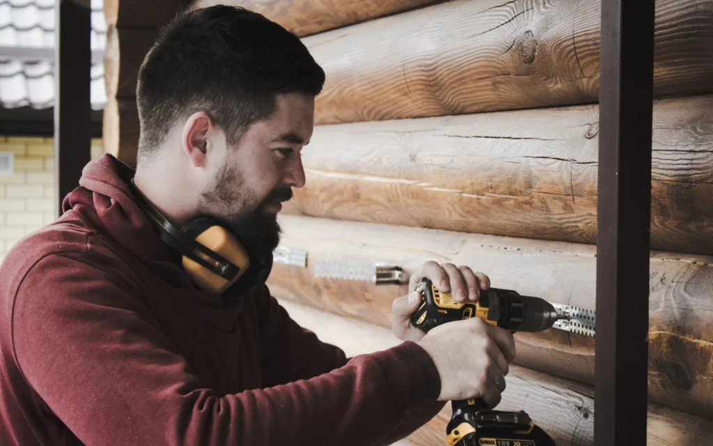 Man in earmuffs using a cordless drill at shoulder height on a timber wall, an overhead task that can aggravate the shoulder